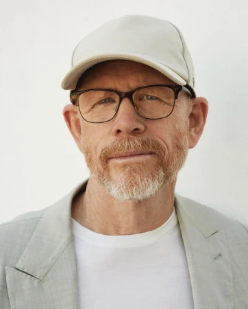 Portrait of Ron Howard wearing glasses, a white baseball cap, white t-shirt, and light grey blazer against a plain white background