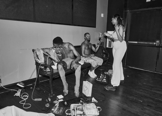 Black and white shot of two shirtless men sitting on a bench, another woman stands nearby with clothing, bags, and styling tools scattered around.