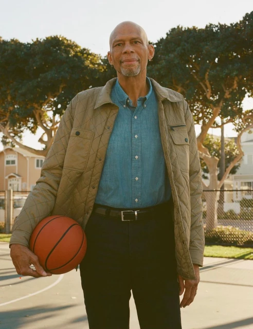 Kareem Abdul-Jabbar stands on an outdoor basketball court holding a basketball, wearing a tan jacket and blue collared shirt.