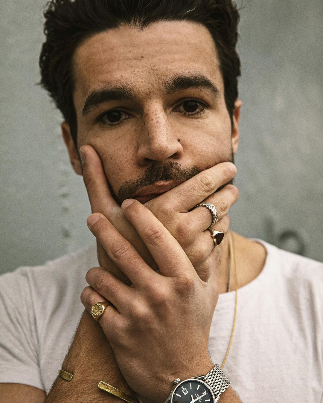 Portrait of a man with dark hair and stubble wearing jewelry with his hands on his face in front of a mottled gray background.