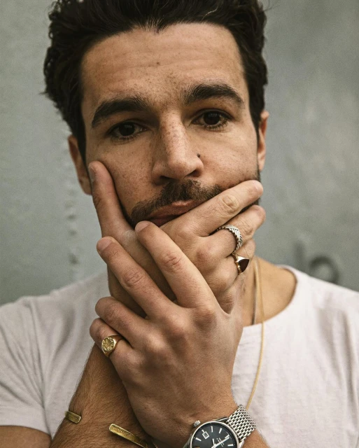 Portrait of a man with dark hair and stubble wearing jewelry with his hands on his face in front of a mottled gray background.