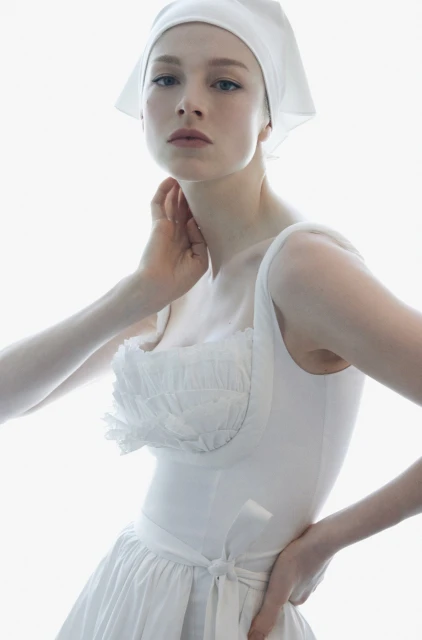 Elegant woman in a white dress and headscarf poses in a brightly lit indoor space, creating a clean and fashionable editorial shot.