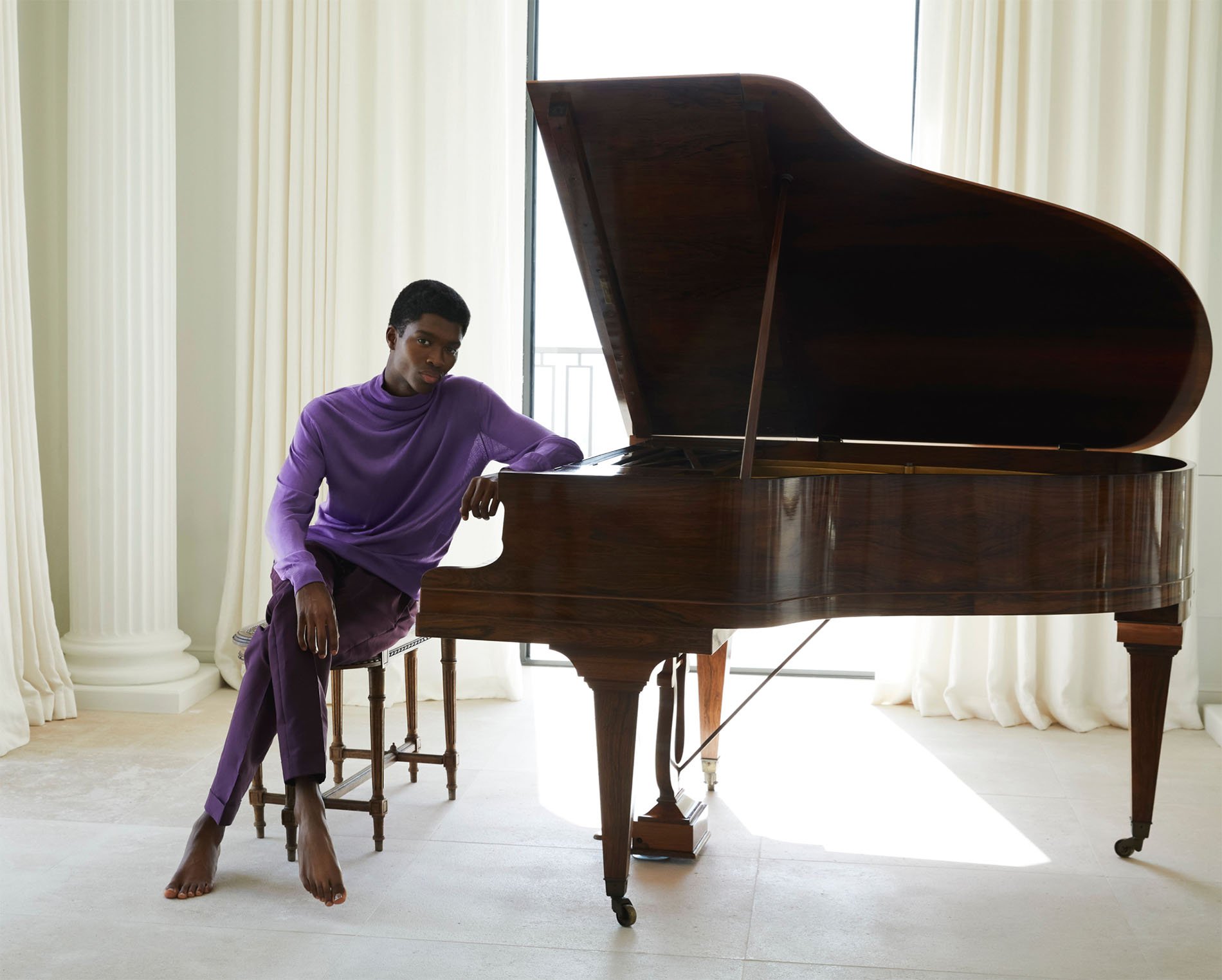 Young Black man in a purple outfit sitting by a grand piano in a light-filled room with flowing white curtains and columns.
