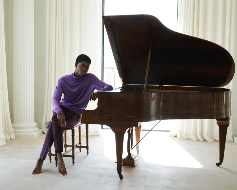 Young Black man in a purple outfit sitting by a grand piano in a light-filled room with flowing white curtains and columns.