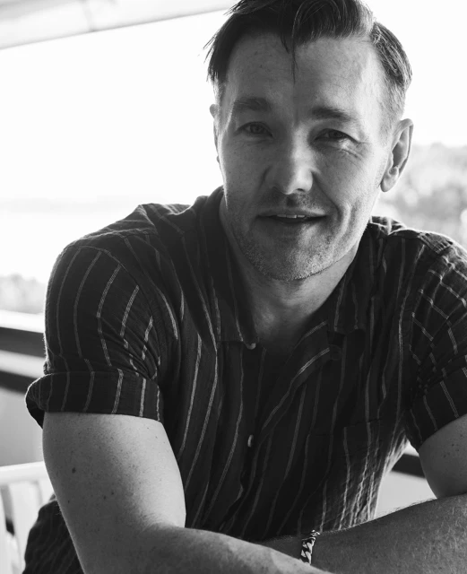 Portrait of adult male in striped shirt with arms folded. Balcony setting with blurred background. Black and white photography.