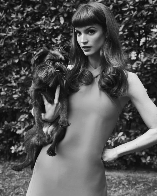 Black and white portrait of an adult woman in a dress holding a Brussels Griffon dog in an outdoor environment with foliage.