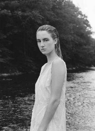 Black and white portrait of a woman in a white dress standing by a river, photographed by Victoria Stevens.