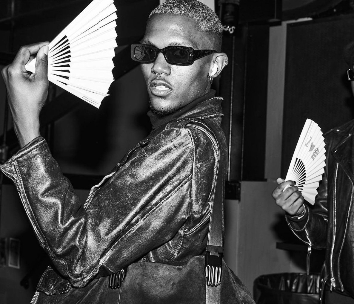 Two people in black and white, one in a leather jacket holding a fan, the other partially visible in the background with a fan.