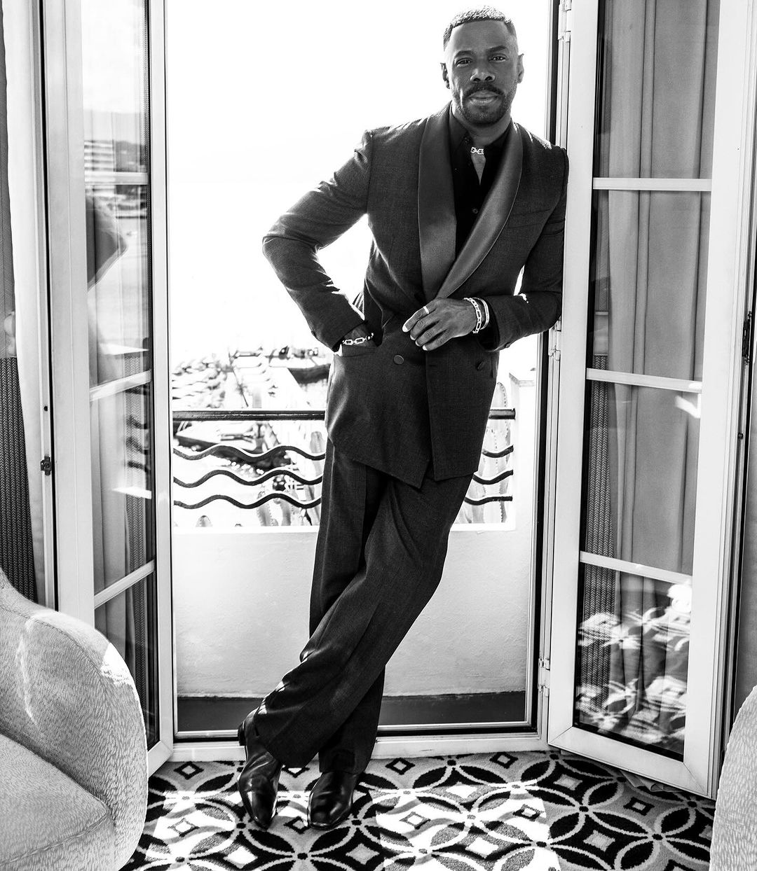 Black and white portrait of an adult male in a dark suit leaning against a doorway on a balcony with a cityscape background.