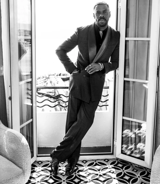 Black and white portrait of an adult male in a dark suit leaning against a doorway on a balcony with a cityscape background.