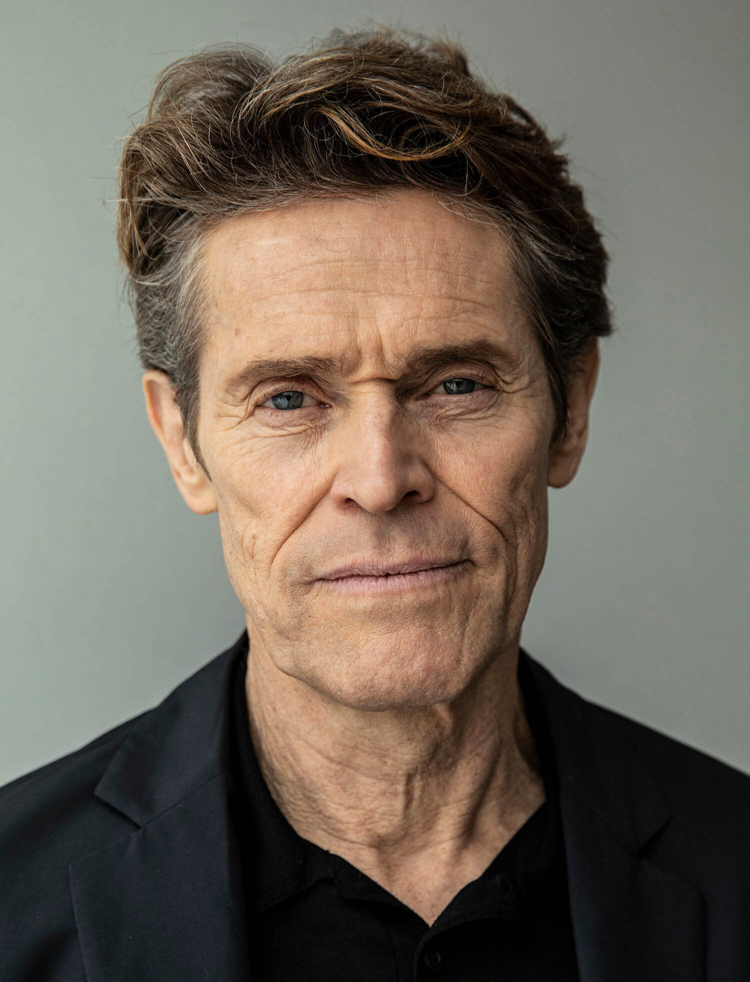 Close-up portrait of Willem Dafoe, an adult male with short brown hair, wearing a black shirt and jacket against a grey background.