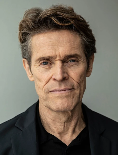 Close-up portrait of Willem Dafoe, an adult male with short brown hair, wearing a black shirt and jacket against a grey background.