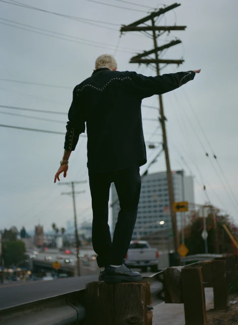 Adult male balancing on roadside barrier against a cloudy sky with utility poles, city buildings, and highway traffic in the background.