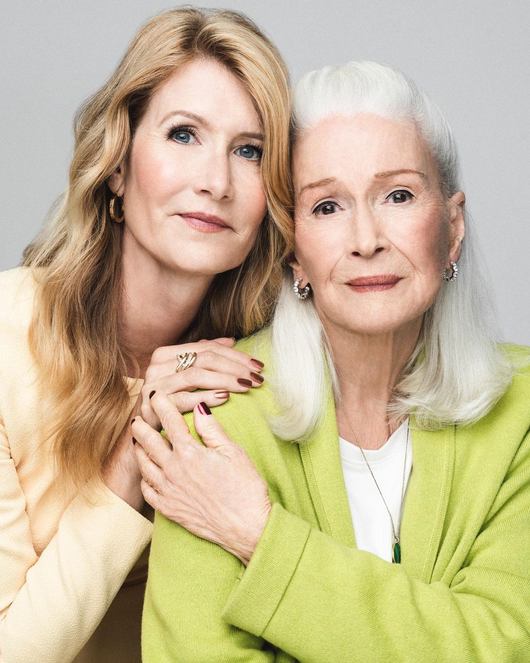 Two women, one with long blonde hair and the other with white hair, pose indoors against a neutral backdrop, exuding a serene, familial warmth.