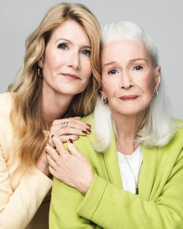 Two women, one with long blonde hair and the other with white hair, pose indoors against a neutral backdrop, exuding a serene, familial warmth.