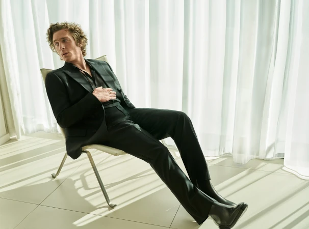 Man with curly hair wearing a dark suit seated in a modern chair before sheer white curtains.