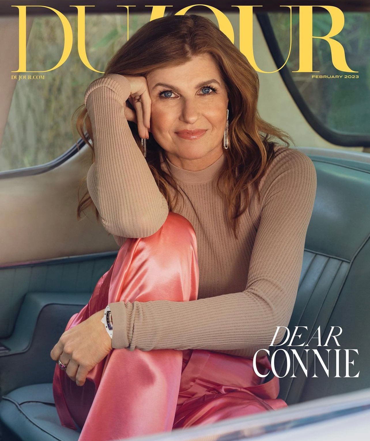 Connie Britton seated inside a vintage car wearing a beige ribbed top and shiny pink trousers, photographed by Victoria Stevens.