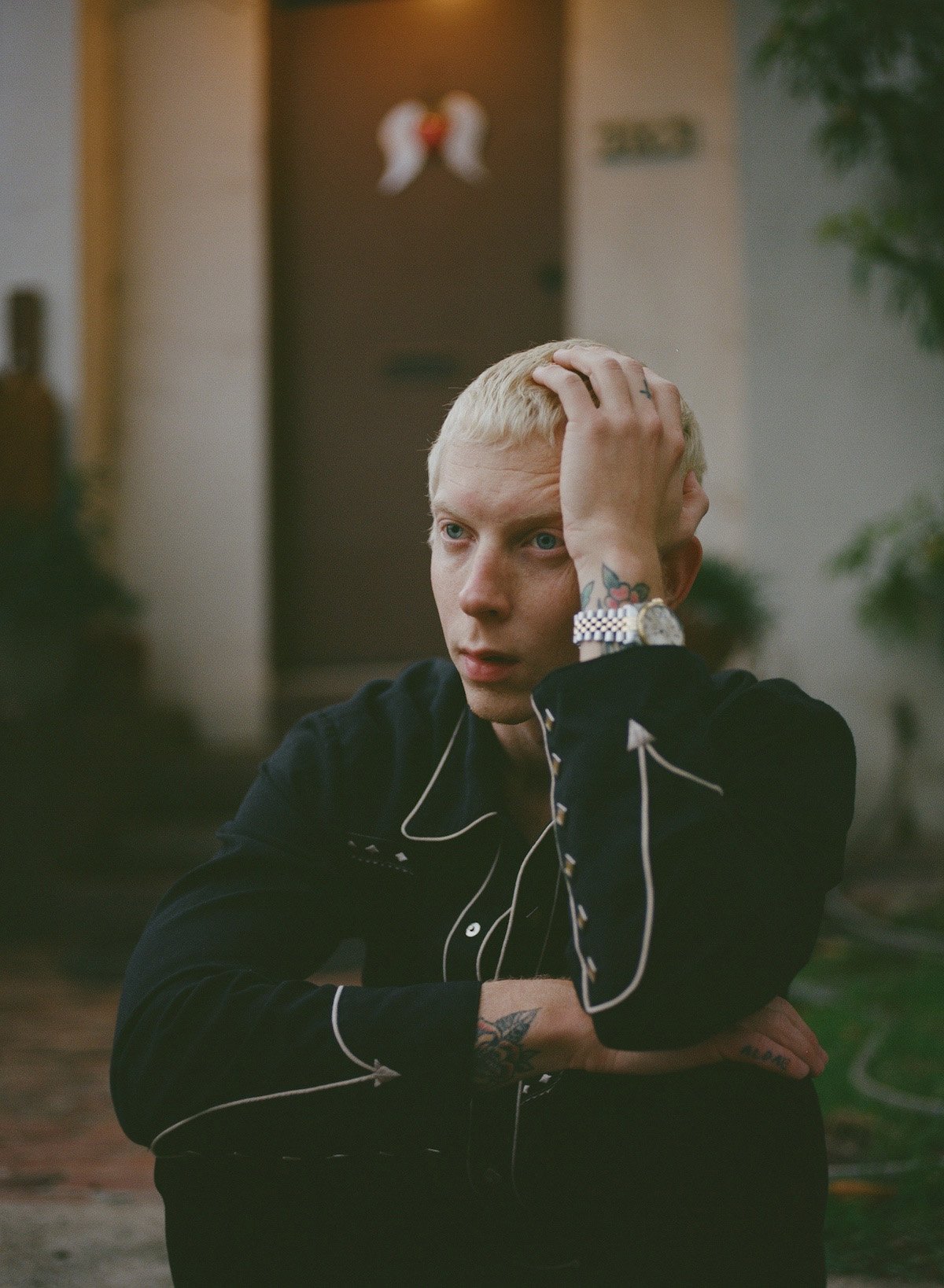 Portrait of a young adult male with blond hair, wearing a black western-style shirt in an outdoor environment in front of a building.