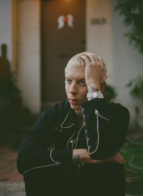 Portrait of a young adult male with blond hair, wearing a black western-style shirt in an outdoor environment in front of a building.