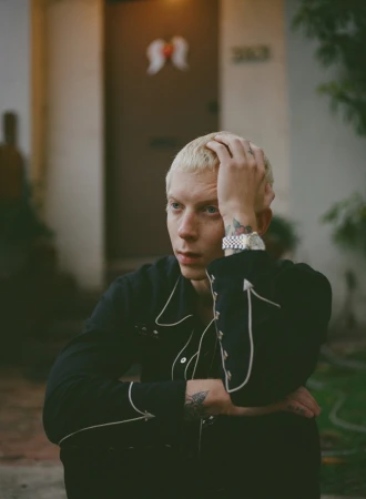 Portrait of a young adult male with blond hair, wearing a black western-style shirt in an outdoor environment in front of a building.