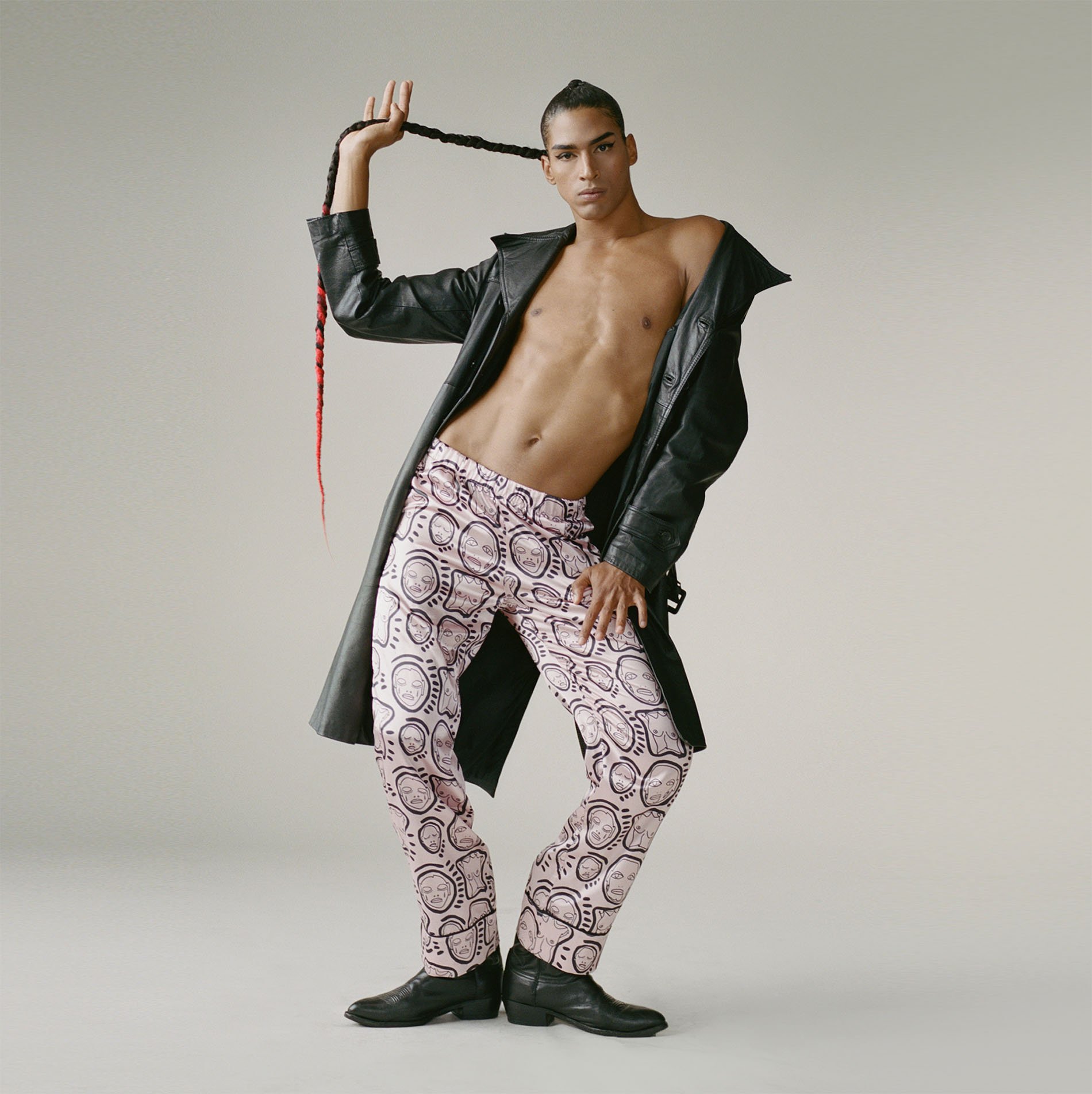 A man in pink patterned pants and a black leather jacket strikes a fashion pose, holding his braided hair up against a grey background.