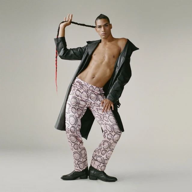 A man in pink patterned pants and a black leather jacket strikes a fashion pose, holding his braided hair up against a grey background.