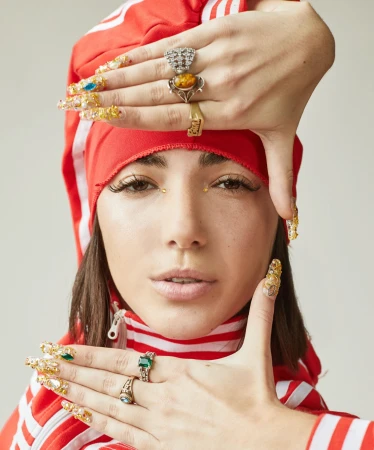 Close-up portrait of a woman in a red track jacket framing her face with hands adorned with ornate rings and jewel-encrusted long nails.