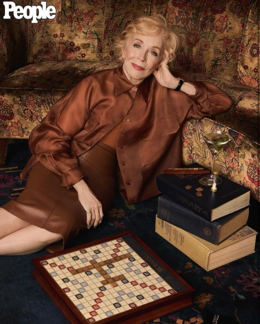 Mature woman with blonde hair poses in a satin top and skirt next to a Scrabble board and martini in a lifestyle setting indoors.