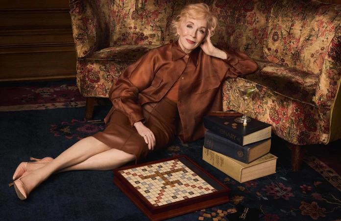 Mature woman in silk blouse poses with vintage board game, books, and cocktail in a moody interior setting with floral couch.