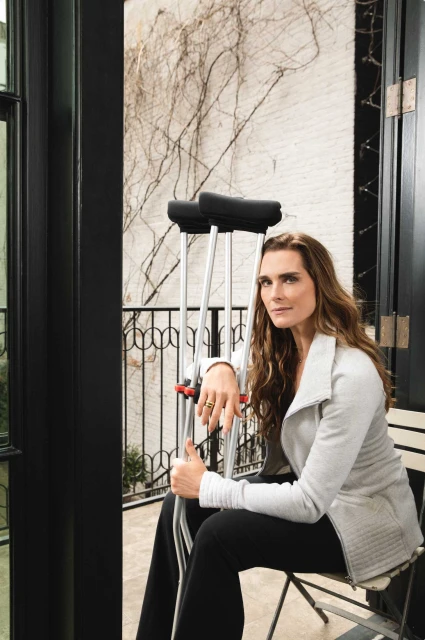 Brooke Shields seated, with crutches, wearing a gray jacket and black pants, gazing at the camera in an indoor setting near a window.