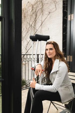 Brooke Shields seated, with crutches, wearing a gray jacket and black pants, gazing at the camera in an indoor setting near a window.