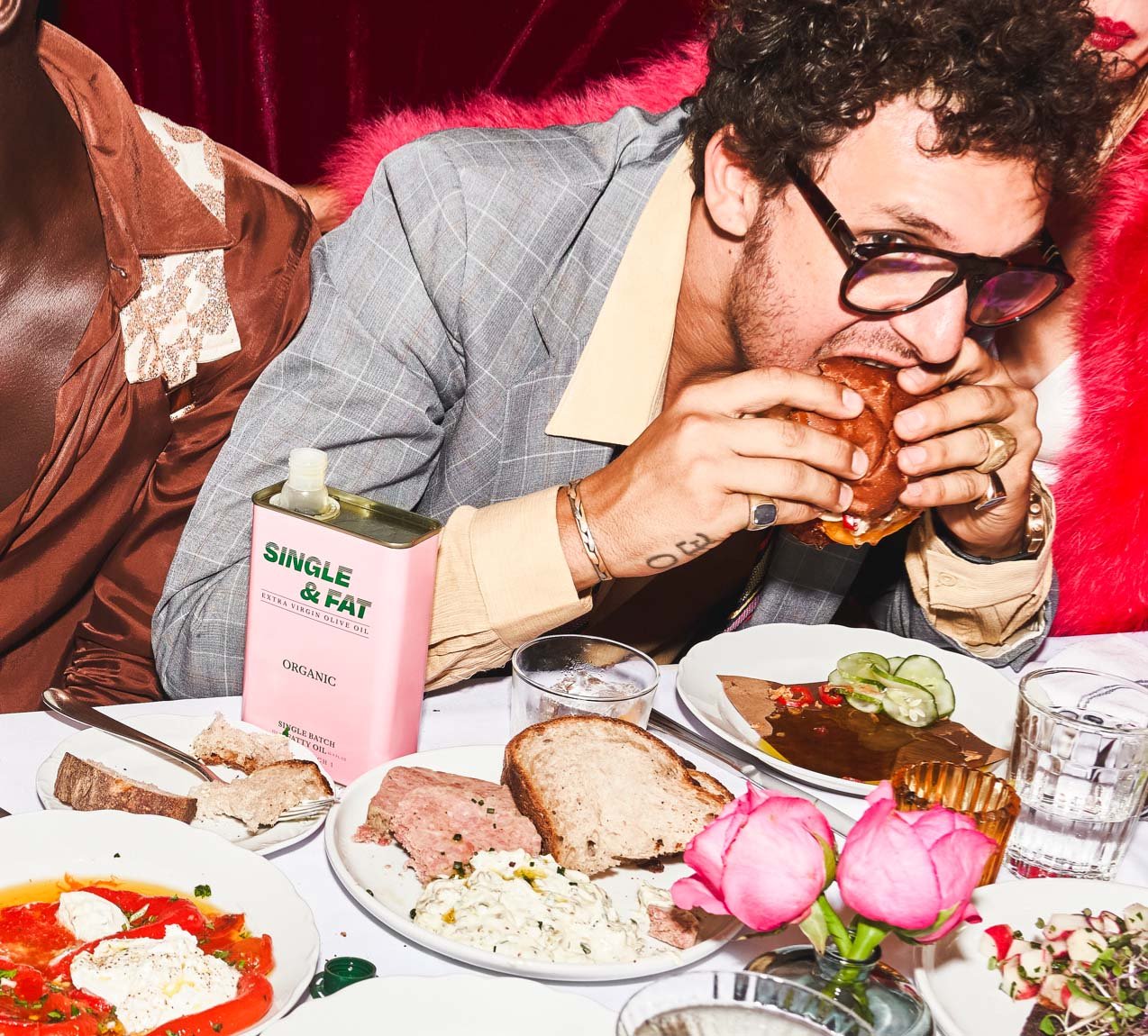 Person in grey suit eating burger at table with salad, bread, canned olive oil, and drinks with a light pink velvet background.