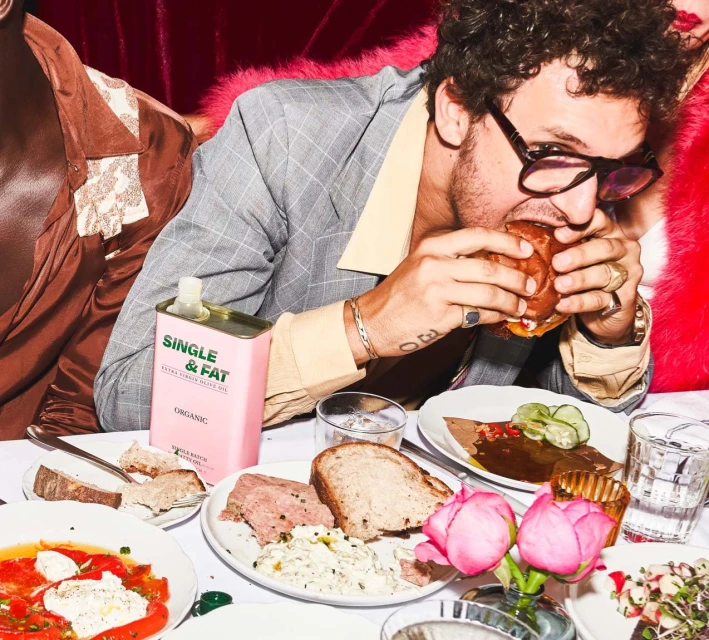 Person in grey suit eating burger at table with salad, bread, canned olive oil, and drinks with a light pink velvet background.