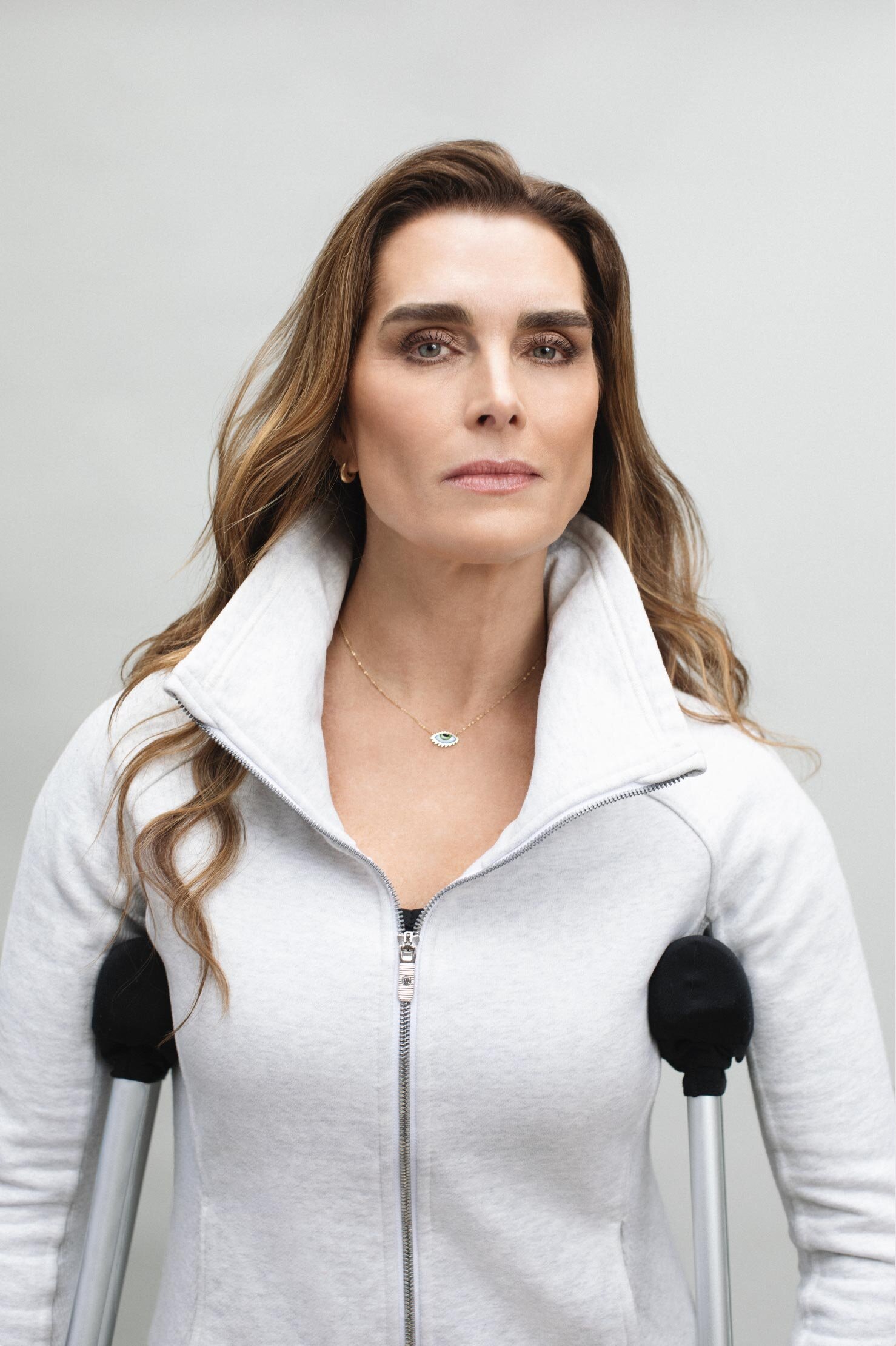 Adult female poses in a medium shot, holding crutches for support, wearing a grey zip up sweater, against a neutral studio backdrop.