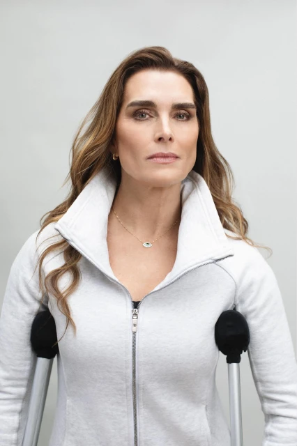 Adult female poses in a medium shot, holding crutches for support, wearing a grey zip up sweater, against a neutral studio backdrop.