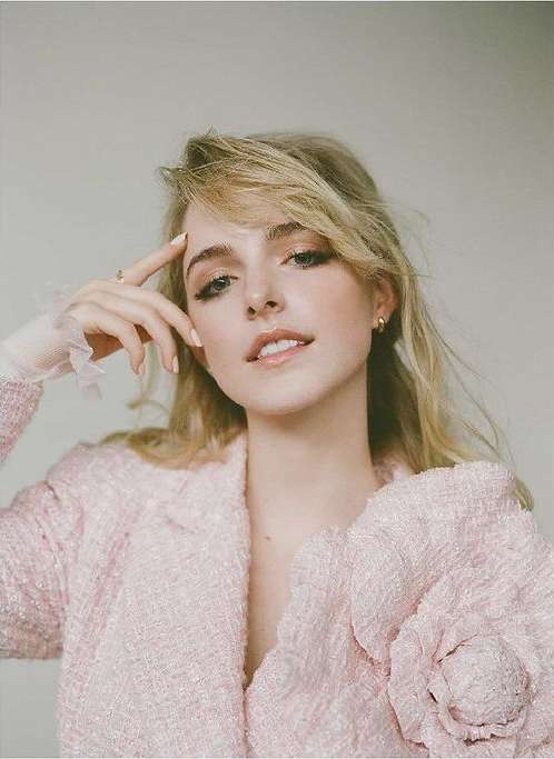 Portrait of a blonde woman with neutral makeup and a pink tweed jacket posing against a plain background in a studio setting.