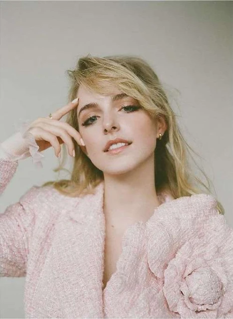 Portrait of a blonde woman with neutral makeup and a pink tweed jacket posing against a plain background in a studio setting.