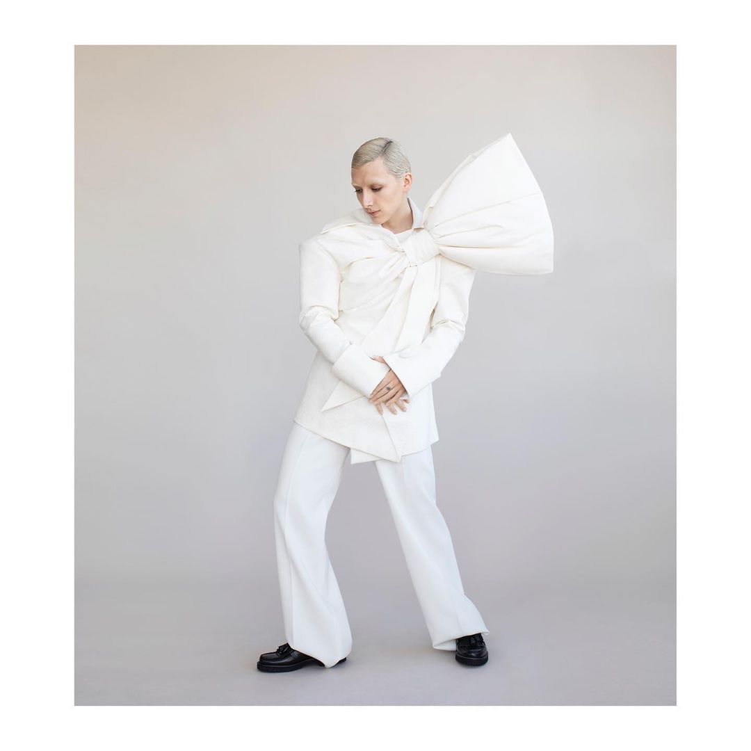A person with short platinum hair poses indoors wearing a white jacket with an oversized bow, matching trousers, and black loafers.