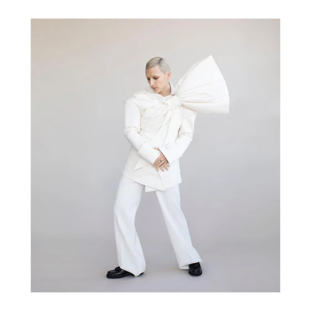 A person with short platinum hair poses indoors wearing a white jacket with an oversized bow, matching trousers, and black loafers.