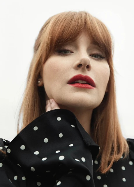Close-up portrait of a red-haired woman with red lipstick and polka dot top, looking directly at the camera against a white background.