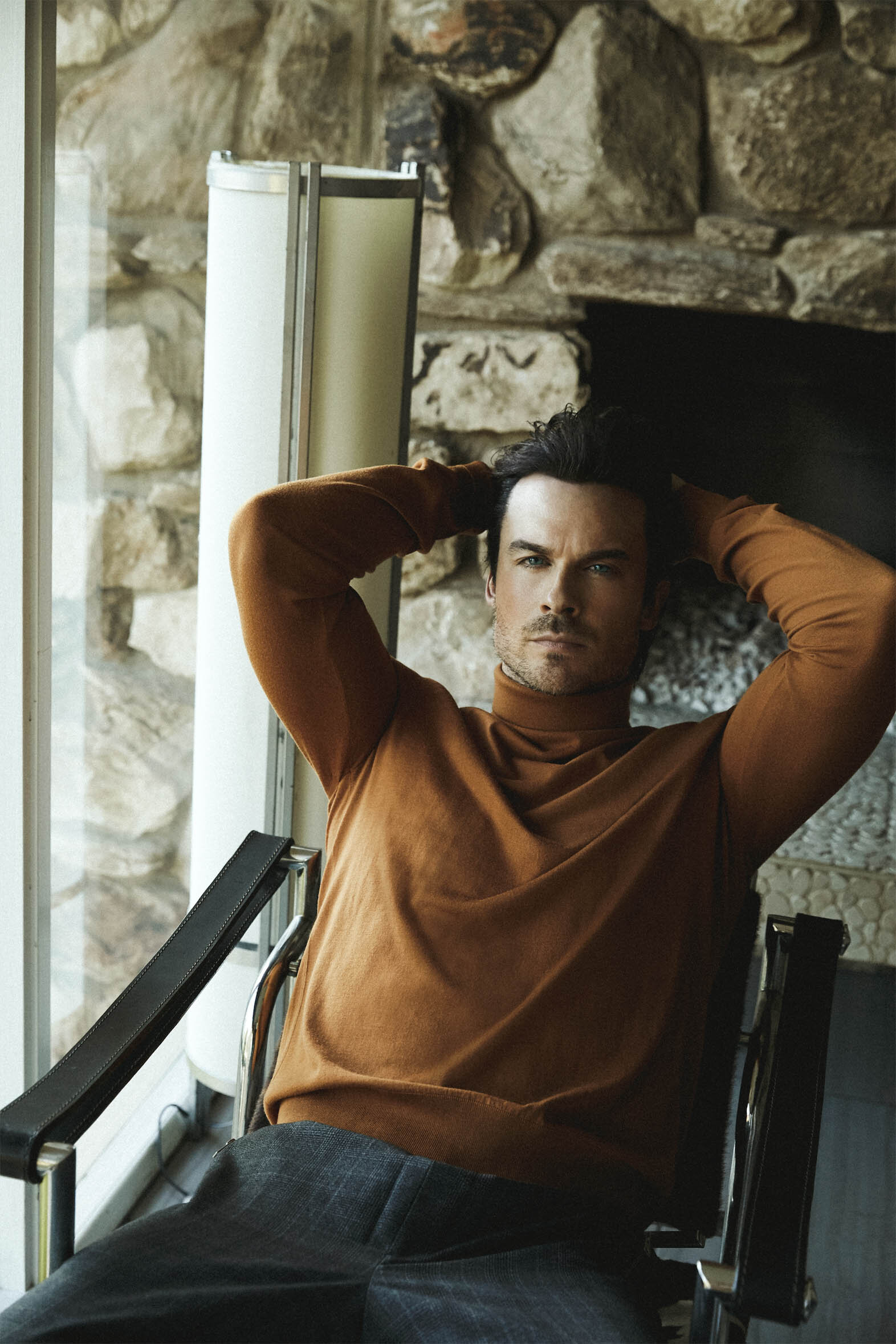 Adult male poses in a rust-colored turtleneck, seated in a chair indoors with stone fireplace background, portrait orientation.