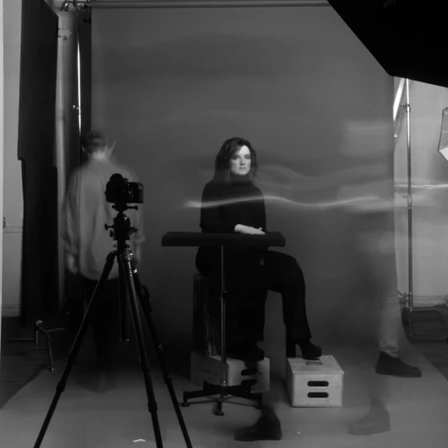 Monochrome portrait of a woman in a black turtleneck, seated in a photo studio during a photo shoot, with visible camera equipment.