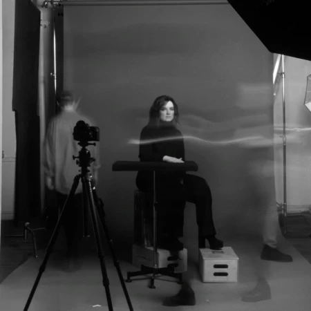 Monochrome portrait of a woman in a black turtleneck, seated in a photo studio during a photo shoot, with visible camera equipment.