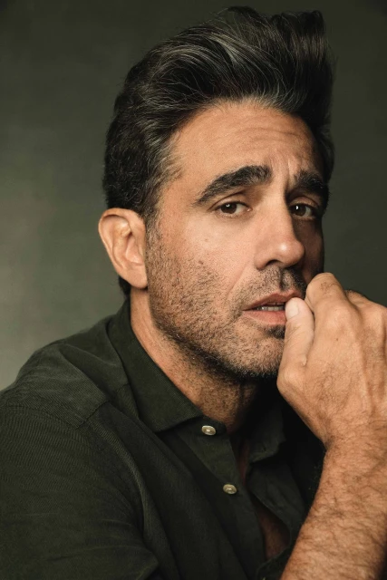 Portrait of an adult male with dark hair and a stubbled beard, wearing a dark green button-down shirt, against a neutral grey backdrop.