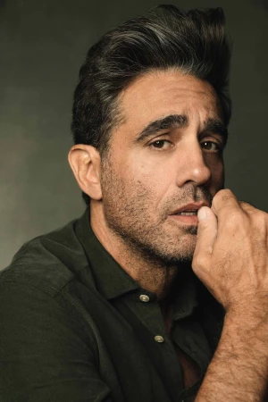 Portrait of an adult male with dark hair and a stubbled beard, wearing a dark green button-down shirt, against a neutral grey backdrop.