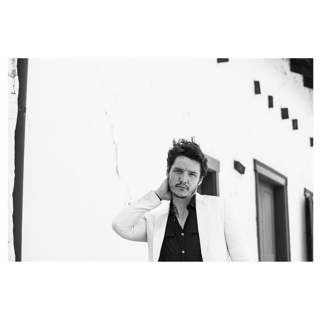 Black and white portrait of adult male outdoors in white jacket with hand on his neck, set against a bright white wall background.