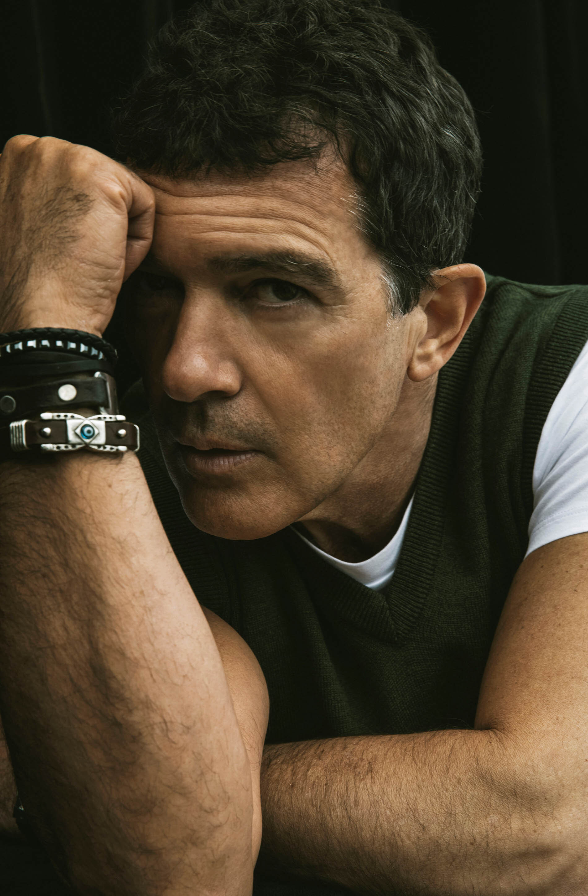 Portrait of adult male resting head on hand wearing an olive green sweater vest with dark hair and bracelets against black background.