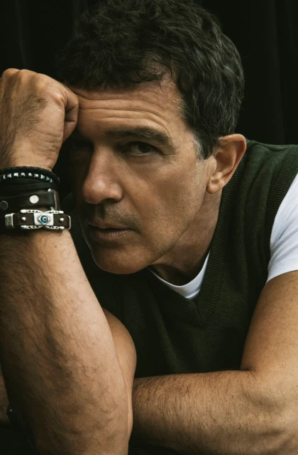 Portrait of adult male resting head on hand wearing an olive green sweater vest with dark hair and bracelets against black background.