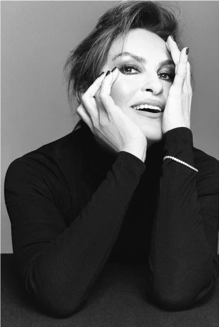 Monochrome portrait of a middle-aged woman with short brown hair, smiling with hands near her face, wearing a black turtleneck sweater.