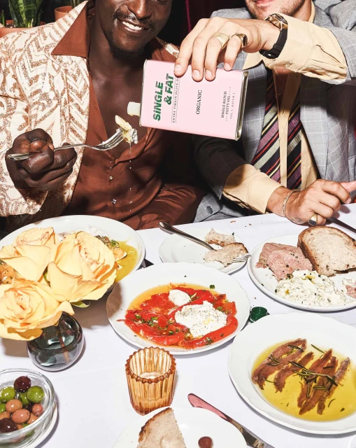 Two men seated at a table laden with various food dishes, one man pours Single & Fat organic olive oil, warm lighting, indoor setting.